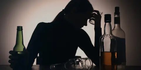 Sylwetka kobiety z kieliszkiem w dłoni, otoczona butelkami. Widoczny problem z przedawkowaniem alkoholu.