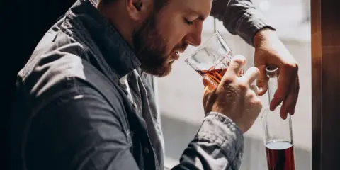 Mężczyzna z brodą, oparty o okno, pije alkohol. W ręku trzyma szklankę z bursztynowym płynem i pustą butelkę. To obraz, jak alkoholik zapije się na śmierć.
