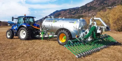 Niebieski traktor ciągnie przyczepę z cysterną do przewozu gnojowicy, wyposażoną w rozdzielacz z wieloma wężami do precyzyjnego nawożenia pól.