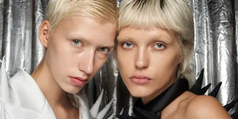 Zwei Models mit Pixie Cut feinem Haar und auffälligen Outfits vor silberner Kulisse.