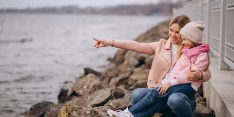 Mama z córką siedzą na kamieniach nad jeziorem. Mama wskazuje coś palcem, a dziewczynka się śmieje. To świetne gąski atrakcje dla dzieci.