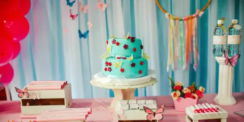 Torta a due piani decorata con farfalle e fiori in pasta di zucchero, su un tavolo rosa con palloncini rossi e caramelle.