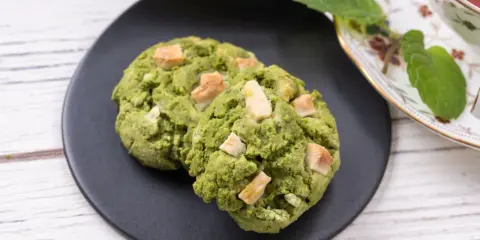 Deux cookies clairs au matcha, parsemés de morceaux de chocolat blanc, sur une assiette noire, à côté d'une tasse de thé et de feuilles de menthe.