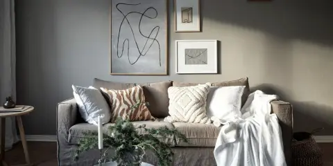 Salon cosy avec un **canapé gris clair** orné de coussins texturés et d'une couverture blanche. Des œuvres d'art abstraites décorent le mur.