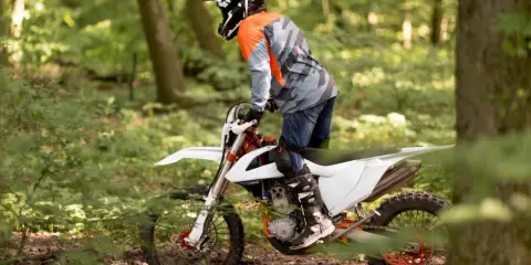 Motocyklista w kasku i pomarańczowej bluzie jedzie na białym crossie przez leśną ścieżkę.