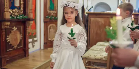 Dziewczynka w białej sukni i wianku trzyma zapaloną świecę. To ważny dzień, w którym klasa przystępuje do komunii.