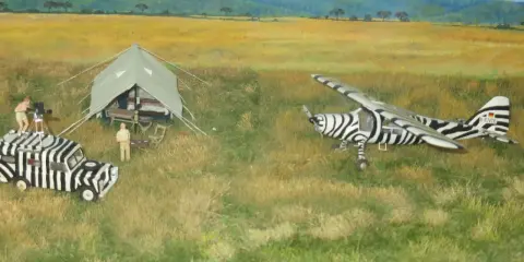 Miniatur-Flugzeug und Auto im Zebradesign in der Savanne. Ein Mann filmt, ein anderer steht neben einem Zelt. do 27.