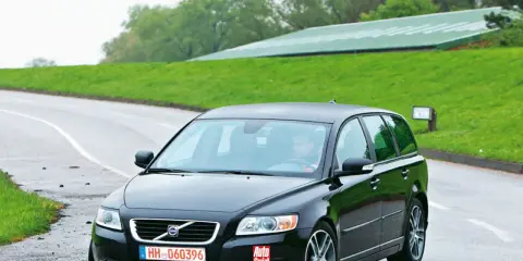 Czarne Volvo V50 2.0D na mokrej drodze, za kierownicą siedzi mężczyzna. W tle zielone wzgórza i budynek.