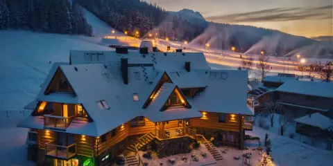 Drewniany pensjonat w śnieżnej scenerii, idealny na zakopane dla początkujących. Oświetlone stoki narciarskie w tle.