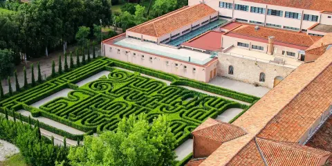 Un giardino con un intricato labirinto borges, circondato da edifici con tetti rossi e alberi verdi.