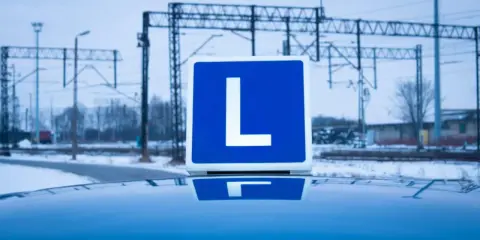 Tablica z literą "L" na dachu samochodu, symbolizująca naukę jazdy i teorię na prawo jazdy. W tle zimowy krajobraz z torami kolejowymi.