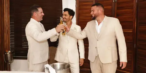 Tres hombres en trajes claros brindan con cerveza. El novio, en el centro, sonríe. Un momento de alegría antes del discurso de boda hermano.