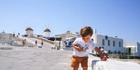Un bambino gioca con una macchinina rossa a Mykonos, con i mulini a vento sullo sfondo. Vacanze a Mykonos con bambini.