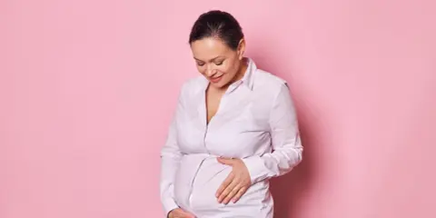 Kobieta w ciąży w wieku 45 lat z uśmiechem na twarzy delikatnie dotyka swojego brzucha.