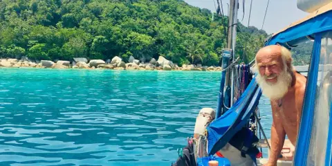 Gangerl Clemens genießt die Fahrt auf seinem Segelboot vor einer üppig grünen Insel mit türkisfarbenem Wasser.