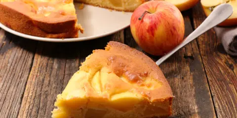 Una fetta di torta di mele senza burro, dorata e soffice, con mele a vista. Accanto, una mela rossa e gialla e altre fette.