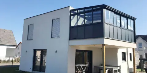 Maison moderne avec une veranda 2 niveaux vitrée, offrant une extension lumineuse. Terrasse en bois en contrebas.