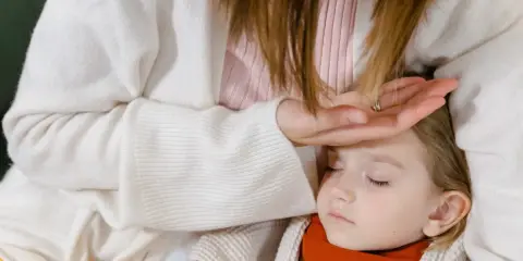 Matka sprawdza czoło chorego dziecka, szukając sposobów, jak zbić gorączkę u dziecka domowym sposobem. Obok leży termometr.