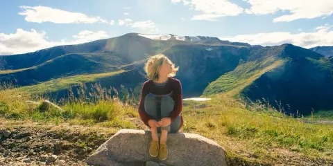 Ragazza seduta su una roccia, con i capelli mossi dal vento, ammira il panorama montano. Un momento di pace mentre camminare nella natura.