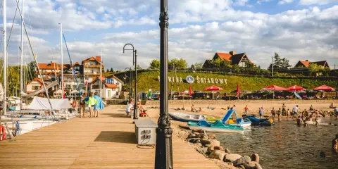 Słoneczna plaża nad jeziorem Śniardwy tętni życiem. Ludzie odpoczywają na piasku, kąpią się w wodzie, a obok cumują żaglówki.