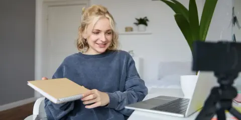 Junge Frau gibt online Nachhilfe, hält ein Notizbuch und lächelt in die Kamera.