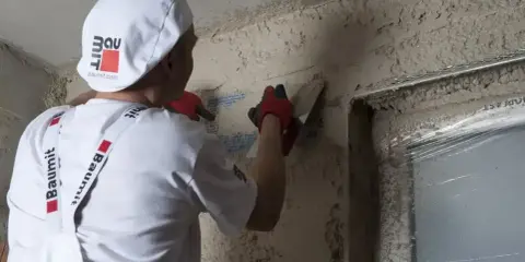 Pracownik w białej czapce i rękawiczkach gąbkuje tynk. Czas na gąbkowanie tynku zależy od jego rodzaju i warunków.