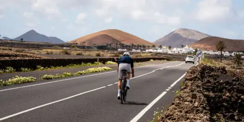 Ein Radfahrer auf einem Rennrad fährt auf Lanzarote durch eine vulkanische Landschaft mit weißen Häusern und Palmen.