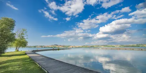 Passeggiata in legno sul **lago di Chiusi**, con cielo azzurro e nuvole bianche che si riflettono sull'acqua calma.