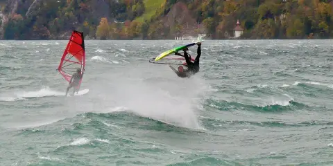 Windsurfer vollführt Sprung am Starnberger See. Ein weiterer Surfer mit rotem Segel im Hintergrund.