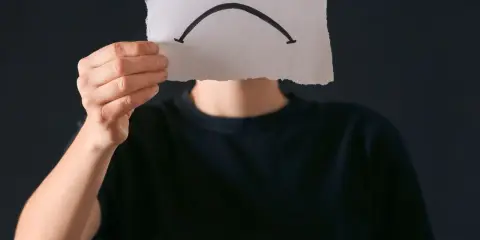 Osoba w czarnej bluzie trzyma kartkę z narysowaną smutną buźką. Czy to anhedonia, co to za uczucie?