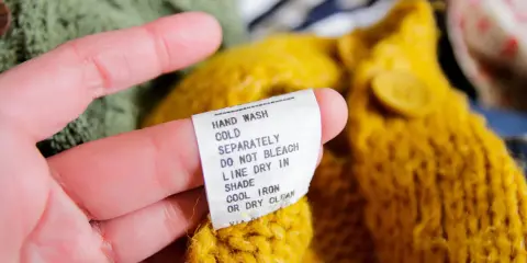 En hand håller i en tvättlapp på en gul stickad tröja. Lappen varnar för att ull kan krympa om den inte sköts rätt.