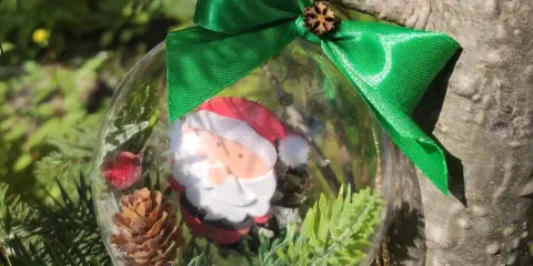 Przezroczysta bombka DIY z Mikołajem, szyszką i gałązkami, przewiązana zieloną wstążką, wisi na choince.