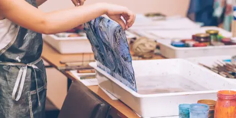Manos con delantal pintan mármol sobre una tela azul y blanca, sumergiéndola en una bandeja con agua.