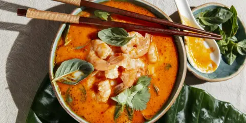 Miseczka zupy tom yum z krewetkami, ozdobiona ziołami, pałeczki i łyżka.