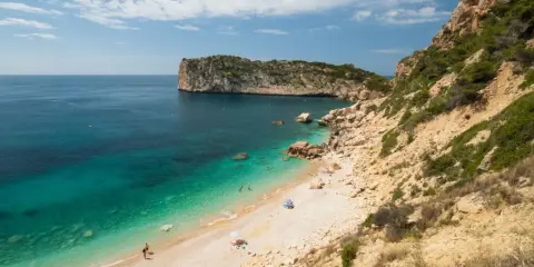 Türkisblaues Wasser trifft auf Kiesstrand in einer malerischen spanischen Atlantikbucht. Felsen und grüne Vegetation säumen die Küste.