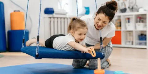 Dziecko na huśtawce podczas terapii integracji sensorycznej co to jest. Terapeuta pomaga dziecku w zabawie.
