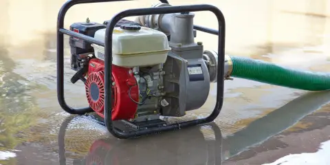 Una motobomba roja y plateada, con una manguera verde, está sumergida en agua turbia, lista para drenar.