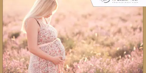 Kobieta w kwiecistej sukience, w 35 tygodniu ciąży, delikatnie dotyka brzucha na tle kwitnącej łąki.