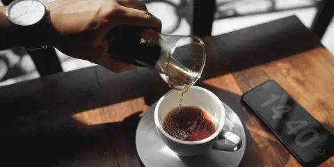 Hand gießt Kaffee in eine Tasse, daneben ein Smartphone mit Uhrzeit.