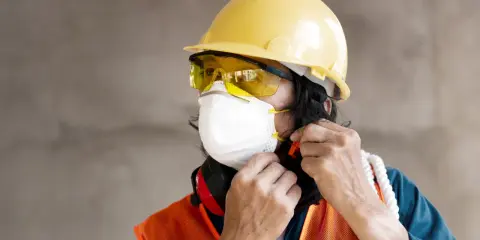 Trabajador ajustando su mascarilla, gafas y epi casco amarillo. Chaleco naranja y protectores auditivos rojos.