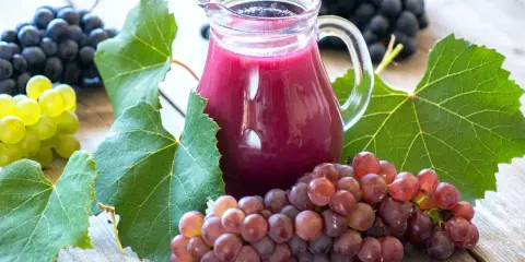 Dzbanek z sokiem z winogron, otoczony kiściami ciemnych i jasnych winogron oraz liśćmi. Odkryj sok z winogron właściwości zdrowotne.