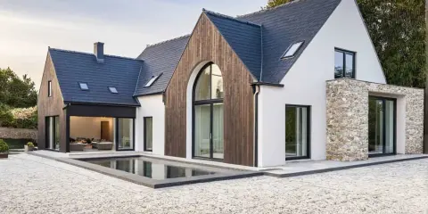 Une maison neuve en pierre et bois, avec une piscine rectangulaire devant.
