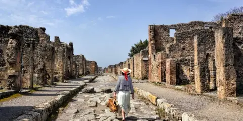 Una donna con cappello di paglia cammina lungo una strada lastricata di Pompei, tra antiche rovine. Un'esperienza unica per scoprire cosa vedere a Pompei.