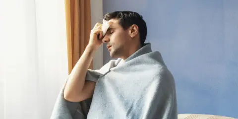Uomo con panno umido sulla fronte, avvolto in una coperta, che sembra soffrire di sudori freddi.