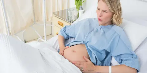 Gravid kvinna i sjukhussäng, med händerna på magen, ser ut att ha värkar. Vattnet har gått och hon väntar på barnet.