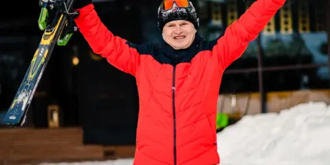 Uśmiechnięty narciarz w czerwonej kurtce trzyma narty, gotowy na stok dla początkujących.