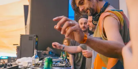 DJ Villa Lobos, con cuffie al collo, mixa musica con passione. Dietro di lui, il pubblico attende il ritmo.