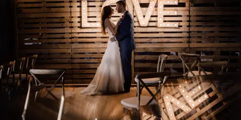 Para nietypowe sesje ślubne, para młoda pozuje przed podświetlanym napisem "LOVE" na tle drewnianych palet.