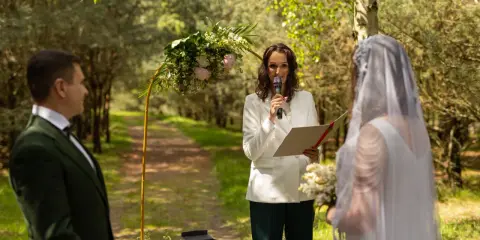 Uroczystość ślubu symbolicznego w lesie. Pan młody w zielonym garniturze, panna młoda w bieli, a prowadząca ceremonię mówi przez mikrofon.