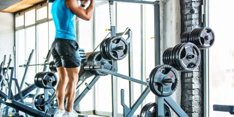 Hombre entrenando en gimnasio, con barra en hombros. Se ven discos de pesas y máquinas.